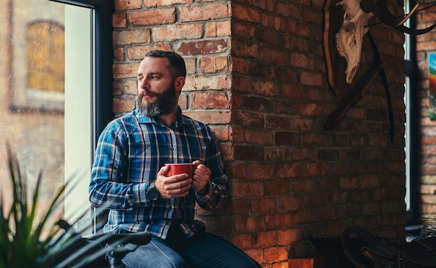 Beau mâle hipster barbu dans une chemise en polaire bleue et un jean tient une tasse de café du matin tout en étant assis sur un rebord de fenêtre dans un studio avec intérieur loft.