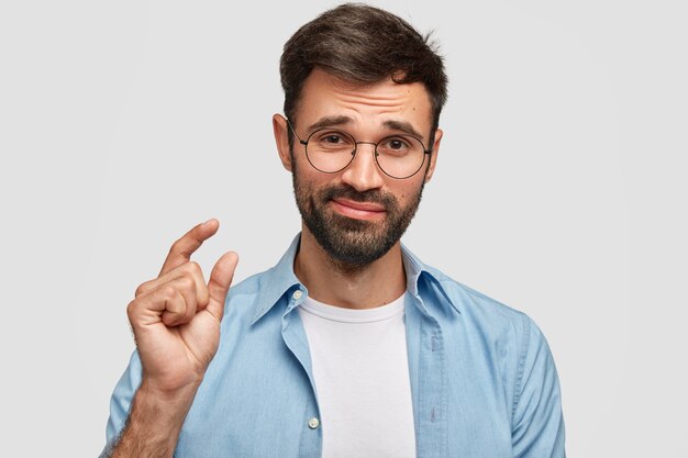 Beau mâle mal rasé avec des cheveux noirs et des poils épais, montre quelque chose de petit avec les mains, vêtu d'une chemise à la mode, isolé sur un mur blanc. Jeune homme montre une petite chose à l'intérieur.