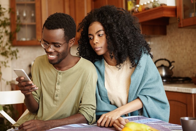 Beau mâle à la peau sombre parcourant les médias sociaux sur smartphone ayant un air heureux, ne remarquant pas sa femme sournoise et suspecte qui l'espionnait. Méfiance, malhonnêteté, tricherie et infidélité