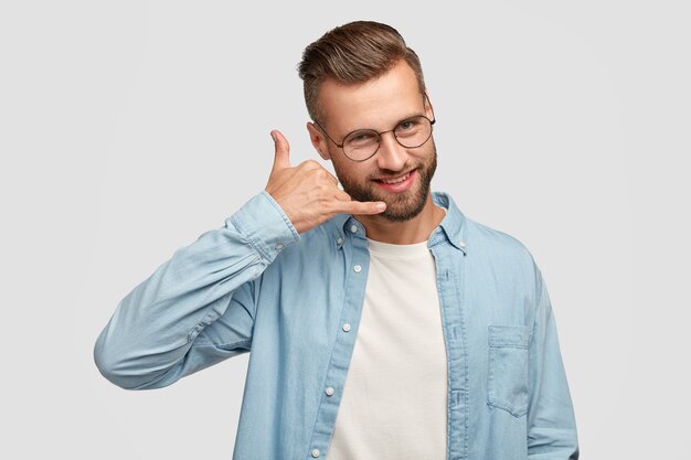 Un beau mâle positif fait un geste téléphonique, fait semblant de parler au téléphone intelligent, a une expression joyeuse, vêtu d'une chemise à la mode, isolé sur un mur blanc. Concept de personnes et de communication