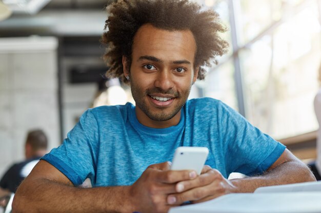 Beau mec afro-américain avec tête de cheveux bouclés assis dans une cafétéria confortable tenant un téléphone intelligent télécharger de la musique à l'aide d'une connexion Internet gratuite à la recherche de sourire heureux et excité