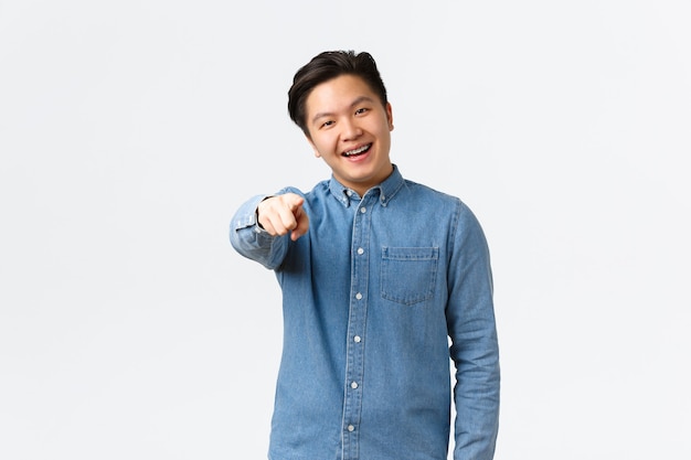 Un beau mec asiatique souriant avec des bretelles faisant le choix, pointant le doigt vers la caméra, choisissant ou choisissant quelque chose, trouvé une personne, debout sur fond blanc, vous félicitant.