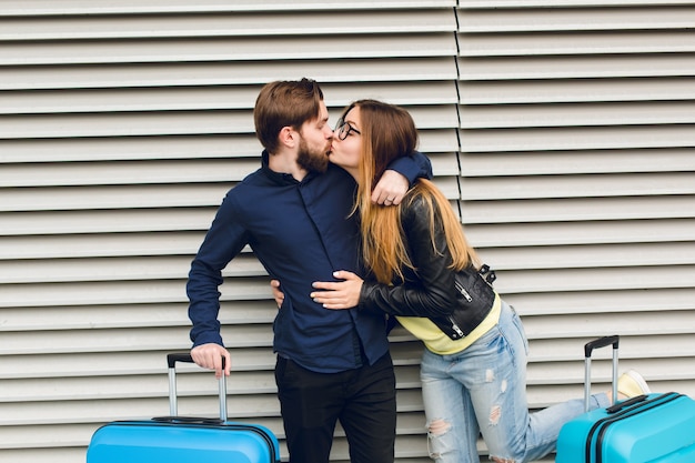 Beau Mec Avec Barbe En Chemise Noire S'embrasse Avec Une Fille Aux Cheveux Longs Sur Fond Rayé Gris Entre Les Valises. Elle Porte Des Lunettes, Un Pull Jaune Et Une Veste Avec Un Jean.