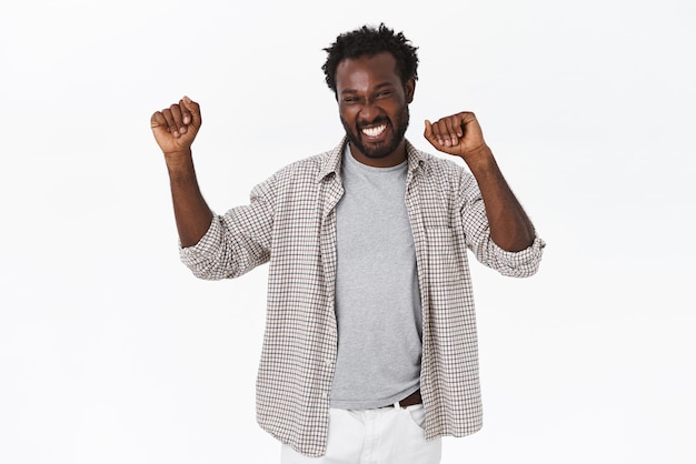 Photo gratuite un beau mec barbu afro-américain heureux et chanceux fait de la danse des champions, levant les mains, souriant joyeusement et triomphant