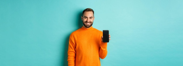 Photo gratuite beau mec barbu en pull orange montrant l'écran du smartphone et souriant montrant la promo en ligne tu