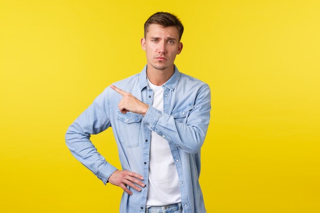 Beau mec blond arrogant et sceptique fronçant les sourcils et n'ayant pas l'air dérangé tout en pointant le coin supérieur gauche vers une bannière promotionnelle moyenne peu amusante, debout sur fond jaune.