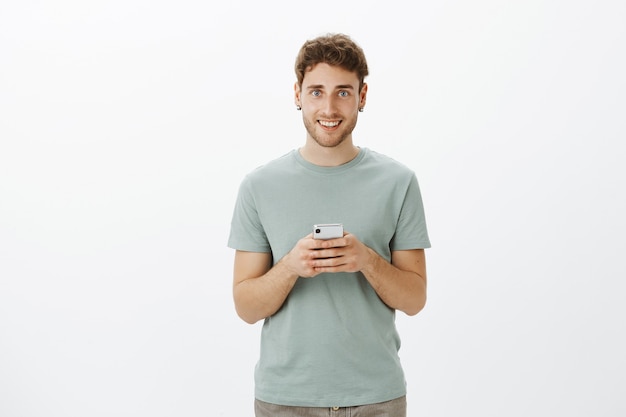 Beau mec blond européen sympathique avec des poils dans des vêtements décontractés, tenant un smartphone, souriant largement, espérant trouver l'amour dans une nouvelle application pour son tout nouvel appareil