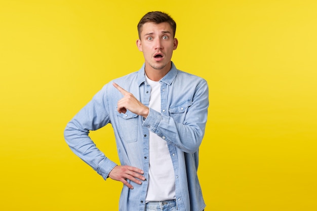 Un beau mec blond inquiet et inquiet à l'air peu sûr et anxieux, pointant le doigt dans le coin supérieur gauche de la caméra fixement alarmé, se sentant effrayé comme debout sur fond jaune.