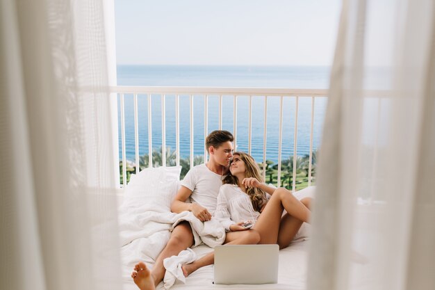 Beau mec brune embrassant sa charmante femme, allongée sur le lit avec un ordinateur portable blanc. Beau jeune couple passer du temps ensemble le matin paresseux, s'embrasser sur un balcon avec vue sur la mer