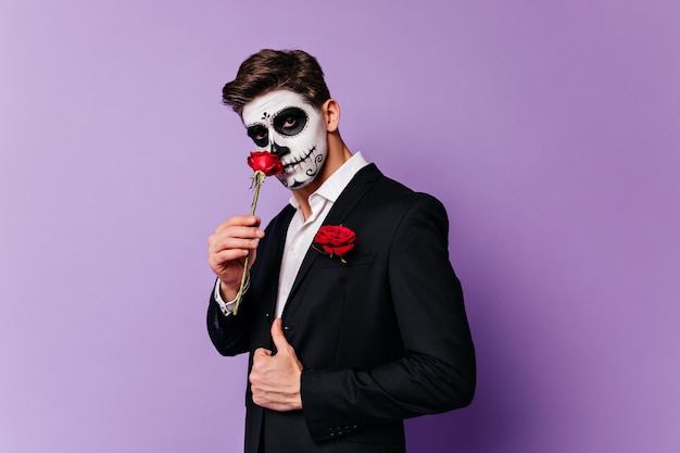 Beau mec caucasien avec maquillage effrayant tenant rose. Photo de Studio d'un modèle masculin bien habillé en tenue de zombie.