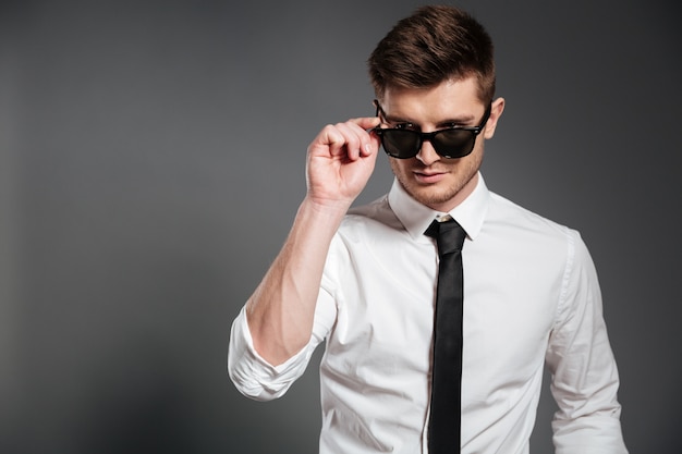 Beau mec en chemise blanche debout et posant avec des lunettes de soleil