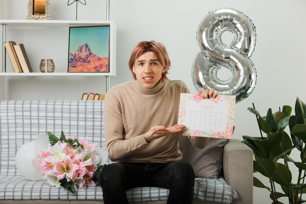 Beau mec confus le jour de la femme heureuse tenant et points avec la main au calendrier assis sur un canapé dans le salon