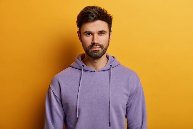 Un beau mec européen barbu sérieux regarde directement, a des poils épais, porte un sweat à capuche violet, habillé d'un sweat à capuche décontracté, pose sur un mur jaune, écoute attentivement les informations.