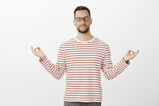 Beau mec européen pratiquant le yoga, portant une tenue et des lunettes à la mode, écartant les mains dans un geste zen et méditant les yeux fermés et un léger sourire, se sentant calme sur un mur gris