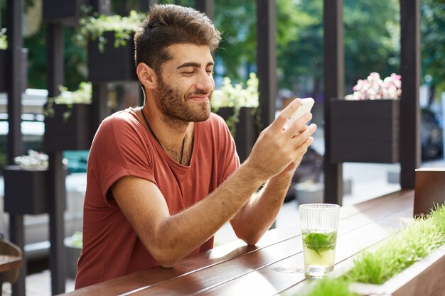 Beau mec heureux assis dans un café, buvant de la limonade et utilisant un téléphone portable, un message texte