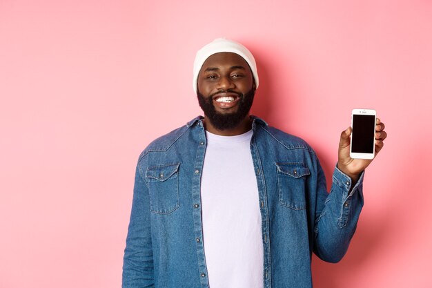 Beau mec hipster en bonnet et chemise en jean souriant, montrant l'écran du téléphone portable avec un visage heureux, introduire l'application, debout sur fond rose