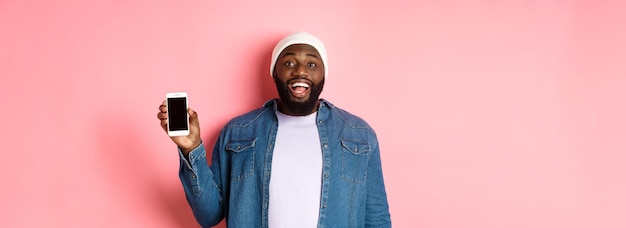 Beau mec hipster en bonnet et chemise en jean souriant montrant l'écran du téléphone portable avec un visage heureux r