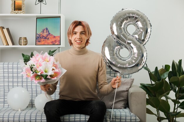 Beau mec le jour de la femme heureuse tenant le ballon numéro huit et le bouquet assis sur le canapé dans le salon