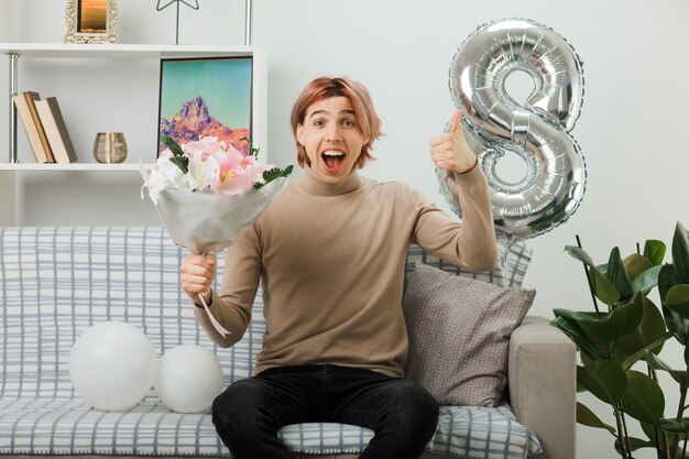 Beau mec le jour de la femme heureuse tenant un bouquet assis sur un canapé dans le salon