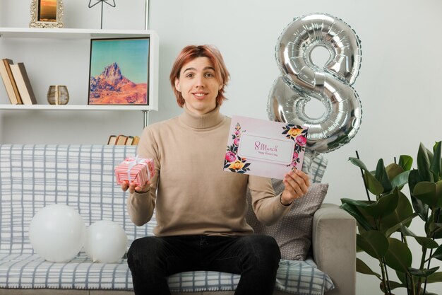 Beau mec le jour de la femme heureuse tenant un cadeau avec carte postale assis sur un canapé dans le salon