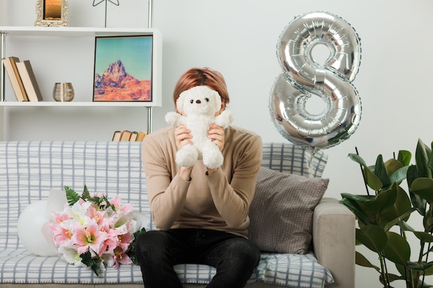 Beau mec le jour de la femme heureuse tenant et visage couvert d'ours en peluche assis sur un canapé dans le salon