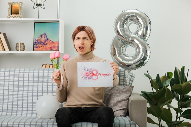 Beau mec mécontent le jour de la femme heureuse tenant des fleurs avec une carte de voeux assis sur un canapé dans le salon