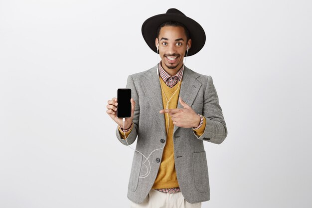 Beau mec noir souriant en costume et chapeau hipster, écoute de la musique dans les écouteurs, pointant le doigt sur l'écran du smartphone