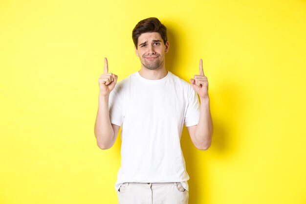 Beau mec non amusé fronçant les sourcils, pointant du doigt quelque chose de mauvais, debout sceptique sur fond jaune.