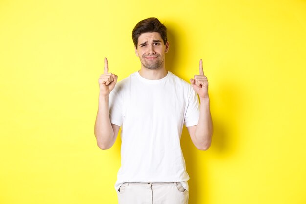 Beau mec sans amusement fronçant les sourcils, pointant du doigt quelque chose de mauvais, debout sceptique sur fond jaune.