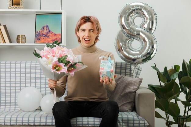 Un beau mec souriant cligna des yeux le jour de la femme heureuse tenant un cadeau avec un bouquet assis sur un canapé dans le salon