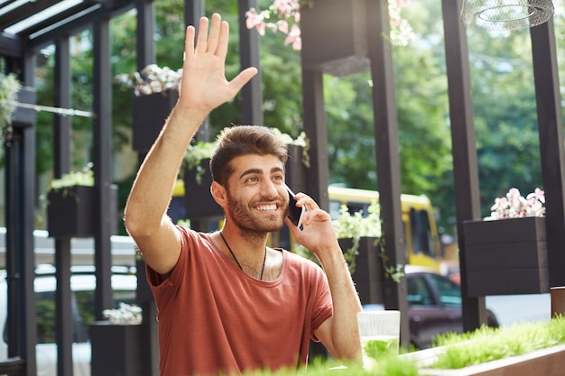 Beau mec souriant parlant au téléphone et agitant la serveuse au café en plein air, demandant la facture