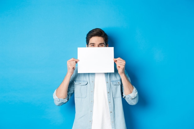 Un beau mec sournois se cachant le visage derrière une feuille de papier vierge pour votre logo, faisant une annonce ou montrant une offre promotionnelle, debout sur fond bleu.