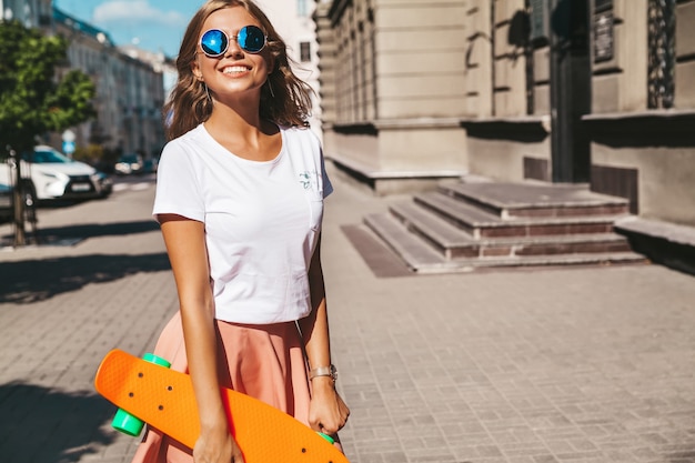 Beau mignon adolescent blond souriant modèle dans des vêtements d'été hipster avec orange penny skateboard posant