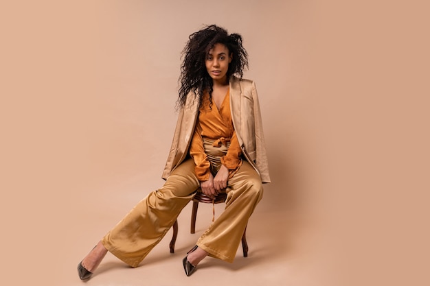 Photo gratuite beau modèle africain avec des cheveux bouclés parfaits dans un élégant chemisier orange et un pantalon en soie assis sur un mur beige de chaise vintage.