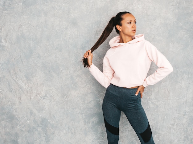 Beau modèle avec un corps bronzé parfait. Femme posant en studio près du mur gris. Tient dans la queue des cheveux à la main