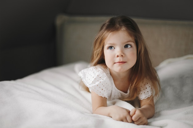 Beau Modèle D'enfant Est Allongé Sur La Couverture Blanche Sur Le Lit Et Regarde Sur Le Côté