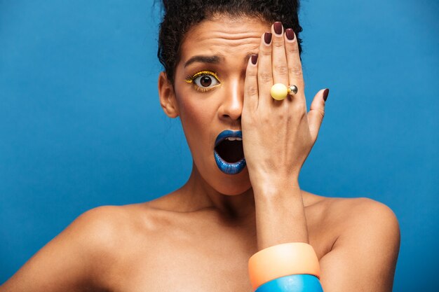 Beau Modèle Féminin Afro Avec Maquillage Coloré Exprimant L'excitation Ou La Surprise Couvrant Un œil Avec La Main, Isolé Sur Le Mur Bleu