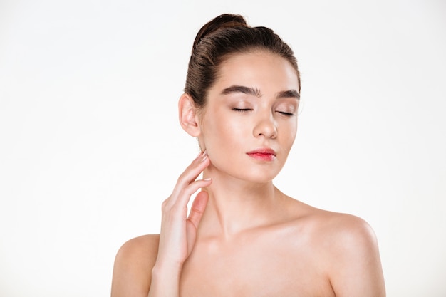 Beau modèle féminin aux cheveux bruns en chignon prenant plaisir avec les yeux fermés tout en faisant des soins de la peau