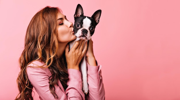 Beau modèle féminin embrassant un drôle de chiot bouledogue Portrait intérieur d'une fille brune raffinée posant sur fond rose avec un chien