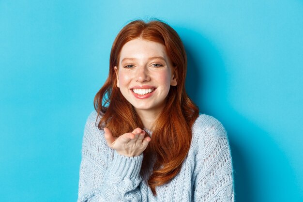 Beau modèle féminin rousse souriant, envoyant un baiser aérien à la caméra, debout sur fond bleu.