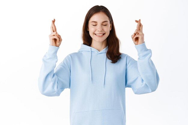 Beau modèle féminin en sweat à capuche faisant son souhait avec les yeux fermés et un large sourire les doigts croisés pour la bonne chance en priant et en espérant que le rêve devienne réalité fond blanc