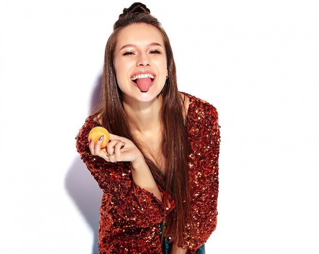 Beau modèle de femme brune hipster souriant caucasien en brillant brillant reflétant la veste élégante d'été et la robe verte isolée. Manger du macaron français et montrer sa langue