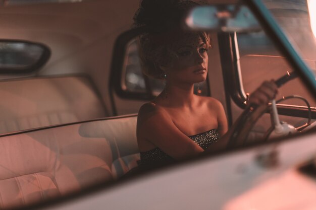 Beau modèle de fille blonde fashion sexy avec maquillage lumineux et coiffure frisée dans un style rétro assis dans une vieille voiture