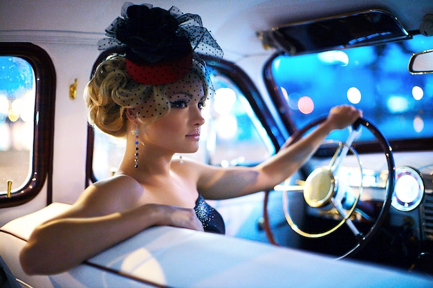 Beau modèle de fille blonde fashion sexy avec maquillage lumineux et coiffure frisée dans un style rétro assis dans une vieille voiture