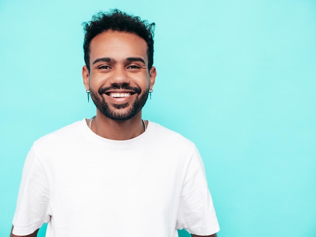 Beau modèle hipster souriant Sexy homme mal rasé habillé en t-shirt d'été blanc et vêtements jeans Mode homme posant près du mur bleu en studio isolé