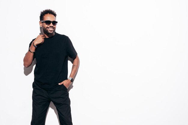 Beau modèle hipster souriant Sexy homme mal rasé habillé en t-shirt d'été noir et vêtements jeans Mode homme posant près d'un mur blanc en studio à la journée ensoleillée Isolé