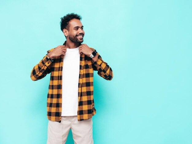 Beau modèle hipster souriant Sexy homme mal rasé vêtu d'une chemise d'été jaune et de vêtements en jean Mode homme posant près du mur bleu en studio Enthousiaste et heureux Isolé