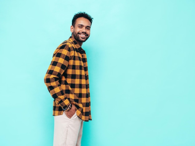 Beau modèle hipster souriant Sexy homme mal rasé vêtu d'une chemise d'été jaune et de vêtements en jean Mode homme posant près du mur bleu en studio Enthousiaste et heureux Isolé