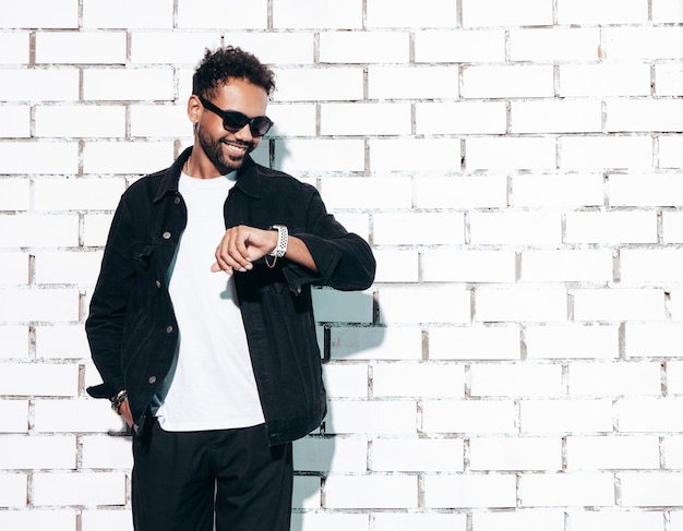 Beau modèle hipster souriant Sexy homme mal rasé vêtu d'une chemise d'été noire et de vêtements en jeans Homme de mode posant près d'un mur de briques blanches en studio à la journée ensoleillée Isolé