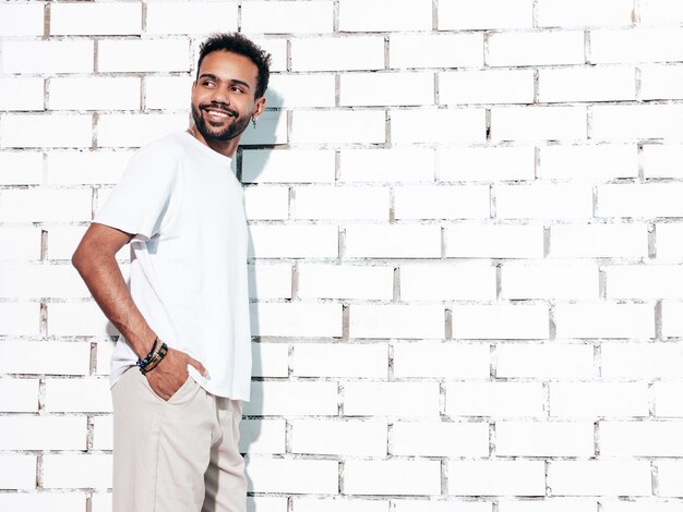 Beau modèle hipster souriant Sexy homme mal rasé vêtu d'un t-shirt d'été blanc et de vêtements en jeans Homme de mode posant près d'un mur de briques blanches en studio par une journée ensoleillée